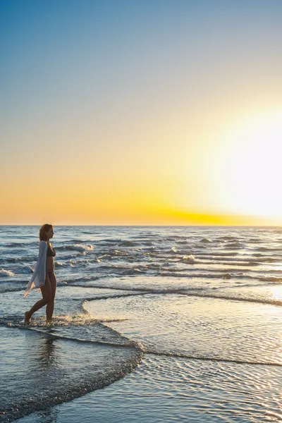 Beautiful mid adult woman in bikini and beach scarf walking in shallow water at sunset